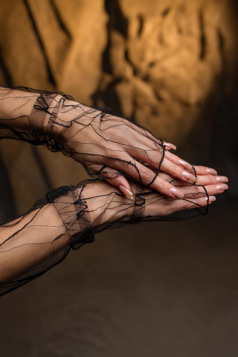 May include: Black, fingerless gloves crafted from fine, sheer lace. The gloves feature a ruffled trim and a delicate, web-like pattern. The hands are posed to showcase the gloves, with long, manicured nails. The backdrop is a warm, neutral shade.