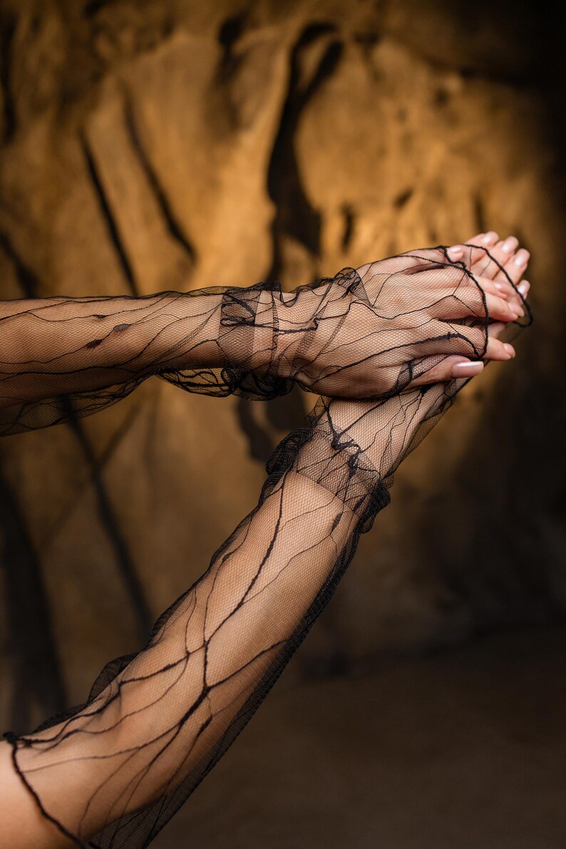 May include: Black, sheer, long gloves with a delicate, web-like pattern. The gloves have a ruffled edge at the wrist. The background is a blurred, dark brown.