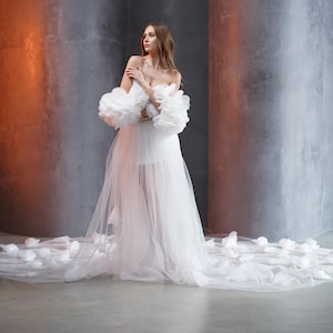 May include: A white tulle gown with a sweetheart neckline and large, ruffled sleeves. The gown has a long train and is decorated with delicate floral appliques.
