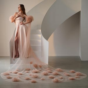 May include: A strapless, blush-colored gown with a high slit and a long, flowing train. The dress features tulle accents on the shoulders and train, adorned with delicate, flower-like embellishments. The model is standing on a white staircase.