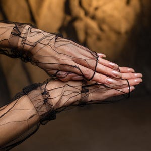 May include: Black, fingerless lace gloves with a delicate, web-like pattern. The gloves feature ruffled edges and extend to the mid-forearm. The background is a warm, neutral tone, highlighting the intricate design of the gloves.