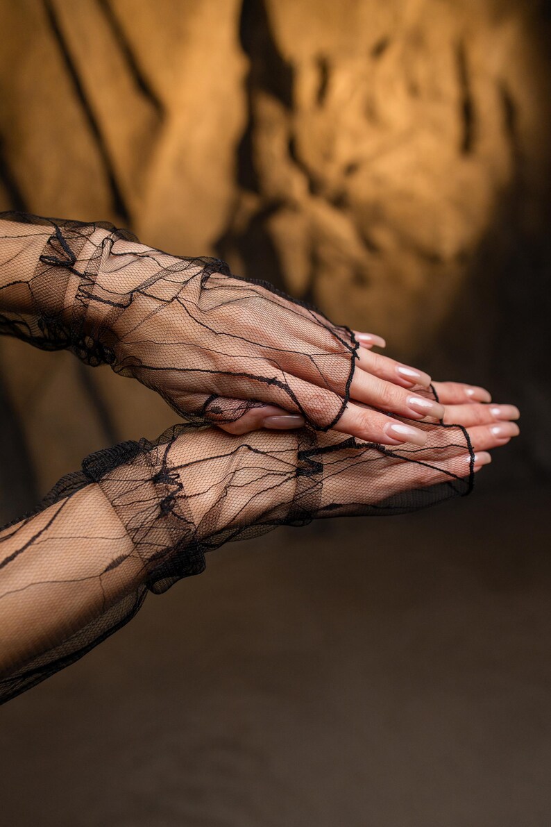 May include: Black, fingerless lace gloves with a delicate, web-like pattern. The gloves feature ruffled edges and extend to the mid-forearm. The background is a warm, neutral tone, highlighting the intricate design of the gloves.