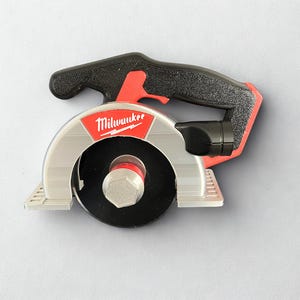 Puede incluir: Una sierra circular de la marca Milwaukee con mango negro, protector de hoja plateado y hoja circular negra. La herramienta presenta una pieza de acento roja cerca del mango y un perno hexagonal central que asegura la hoja en su posici&oacute;n.