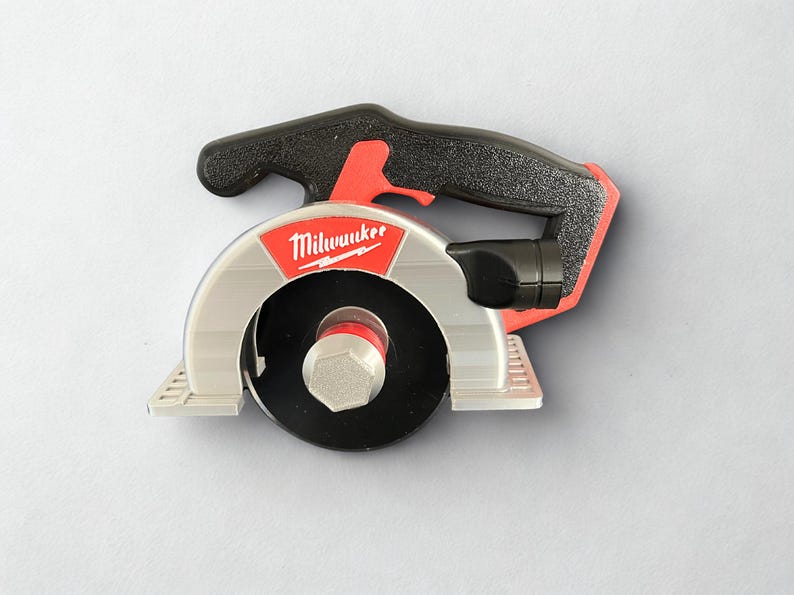 Puede incluir: Una sierra circular de la marca Milwaukee con mango negro, protector de hoja plateado y hoja circular negra. La herramienta presenta una pieza de acento roja cerca del mango y un perno hexagonal central que asegura la hoja en su posici&oacute;n.