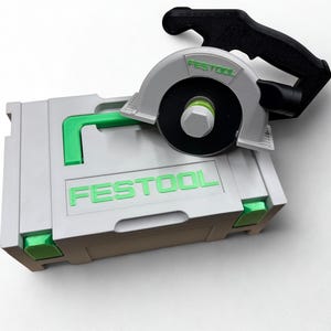 Puede incluir: Un malet&iacute;n de almacenamiento de pl&aacute;stico gris claro con detalles en verde brillante y la palabra FESTOOL impresa en verde. Sobre el malet&iacute;n descansa un modelo de sierra circular en miniatura con protector gris claro, mango negro y logo verde.