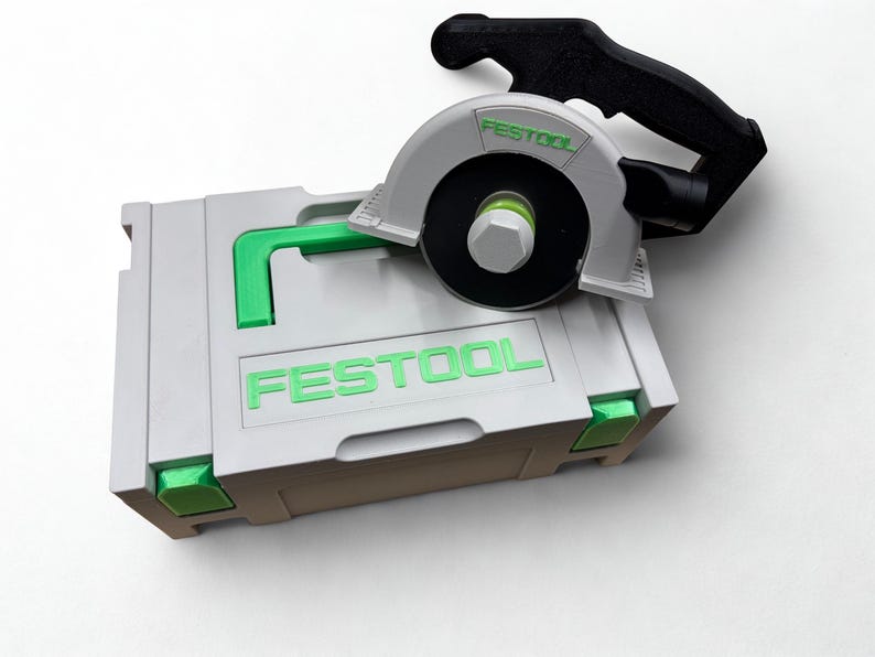 Puede incluir: Un malet&iacute;n de almacenamiento de pl&aacute;stico gris claro con detalles en verde brillante y la palabra FESTOOL impresa en verde. Sobre el malet&iacute;n descansa un modelo de sierra circular en miniatura con protector gris claro, mango negro y logo verde.