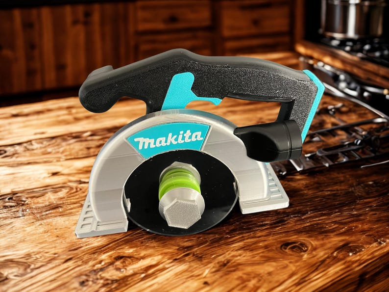 Puede incluir: Una r&eacute;plica de herramienta de sierra circular de la marca Makita con carcasa plateada, mango negro con detalles en turquesa y una zona central de hoja circular negra con un perno hexagonal de color verde lima y plata, con textura en capas.