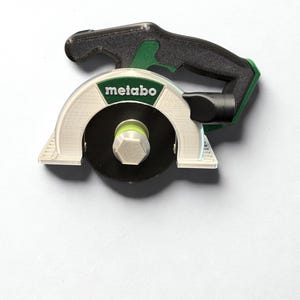 Puede incluir: Un modelo en miniatura de sierra circular impreso en 3D con un mango negro, un protector de hoja blanco con el logotipo verde de Metabo y una hoja circular negra con una tuerca hexagonal central. La herramienta muestra l&iacute;neas de capa.