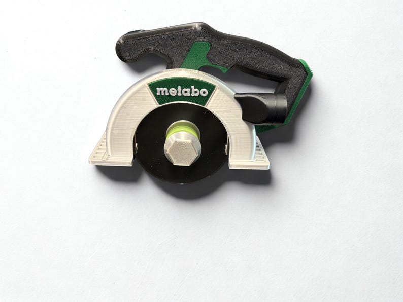 Puede incluir: Un modelo en miniatura de sierra circular impreso en 3D con un mango negro, un protector de hoja blanco con el logotipo verde de Metabo y una hoja circular negra con una tuerca hexagonal central. La herramienta muestra l&iacute;neas de capa.