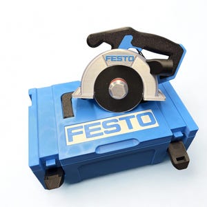 Puede incluir: Una sierra circular Festo con mango negro y protector de hoja plateado sobre un malet&iacute;n de transporte de pl&aacute;stico azul. El malet&iacute;n presenta el logotipo de Festo en texto blanco sobre fondo azul, y la sierra tambi&eacute;n muestra la marca en su protector.