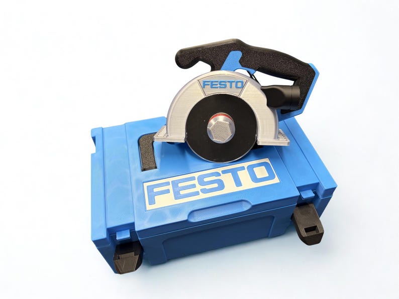 Puede incluir: Una sierra circular Festo con mango negro y protector de hoja plateado sobre un malet&iacute;n de transporte de pl&aacute;stico azul. El malet&iacute;n presenta el logotipo de Festo en texto blanco sobre fondo azul, y la sierra tambi&eacute;n muestra la marca en su protector.