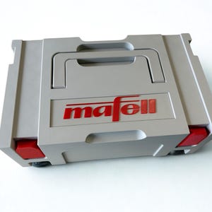 Puede incluir: Un malet&iacute;n de transporte rectangular de pl&aacute;stico gris claro con un asa superior y cierres rojos en los laterales. El nombre de la marca mafell est&aacute; impreso en letras rojas en el centro de la parte frontal. El malet&iacute;n presenta un dise&ntilde;o estructurado.