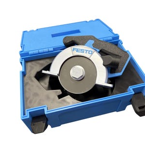 Puede incluir: Un accesorio de hoja de sierra circular Festo con carcasa de metal plateado y un perno central, alojado dentro de un malet&iacute;n de transporte de pl&aacute;stico azul con inserciones de espuma negra. La marca Festo est&aacute; impresa en azul en la parte superior.
