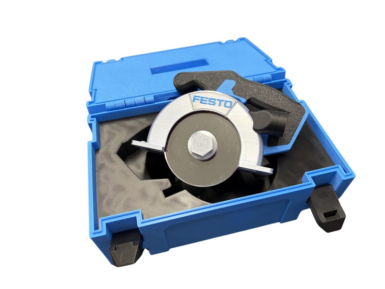 Puede incluir: Un accesorio de hoja de sierra circular Festo con carcasa de metal plateado y un perno central, alojado dentro de un malet&iacute;n de transporte de pl&aacute;stico azul con inserciones de espuma negra. La marca Festo est&aacute; impresa en azul en la parte superior.