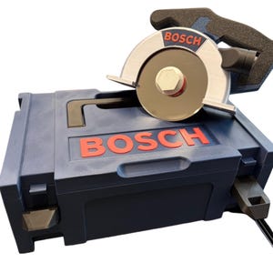 Puede incluir: Una sierra circular Bosch de color azul con una hoja de metal plateado y un mango negro, apoyada sobre un malet&iacute;n de transporte de pl&aacute;stico Bosch azul a juego. El nombre de la marca Bosch aparece en letras rojas en la herramienta y el estuche.
