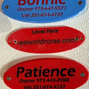 Puede incluir: Tres etiquetas ovaladas con texto. La etiqueta superior es azul y dice "Bonnie" con un número de teléfono y un número de veterinario. La etiqueta del medio es roja y dice "Level Here" y "realworldhorse.com". La etiqueta inferior es roja y dice "Patience" con un número de teléfono y un número de veterinario.