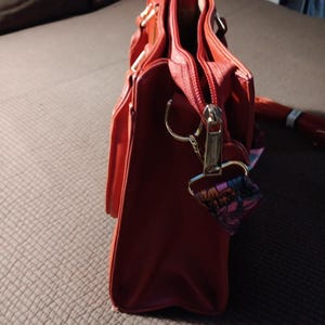 K&ouml;nnte beinhalten: Eine rote Ledertasche mit Rei&szlig;verschluss und goldfarbenen Beschl&auml;gen. Die Tasche hat einen Henkel oben und einen abnehmbaren Schultergurt.