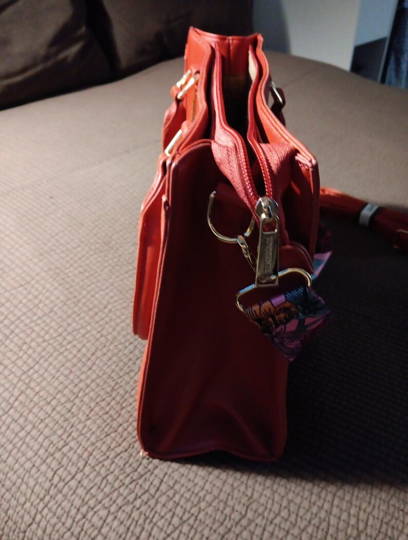 K&ouml;nnte beinhalten: Eine rote Ledertasche mit Rei&szlig;verschluss und goldfarbenen Beschl&auml;gen. Die Tasche hat einen Henkel oben und einen abnehmbaren Schultergurt.