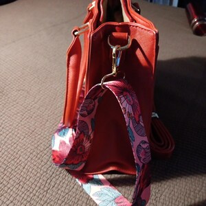 K&ouml;nnte beinhalten: Eine rote Handtasche mit einem floralen Riemen. Die Tasche hat einen goldenen Rei&szlig;verschluss und einen goldenen Griff.