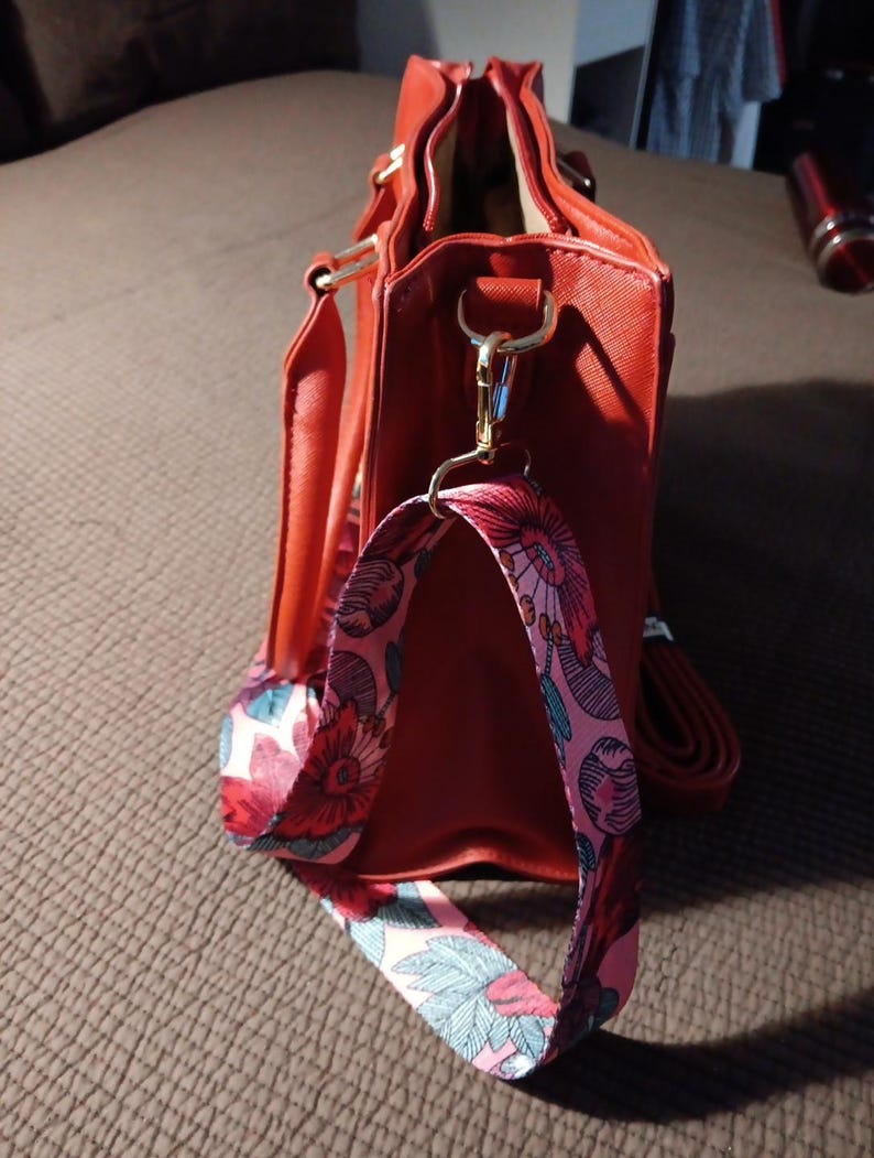 K&ouml;nnte beinhalten: Eine rote Handtasche mit einem floralen Riemen. Die Tasche hat einen goldenen Rei&szlig;verschluss und einen goldenen Griff.
