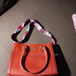 K&ouml;nnte beinhalten: Eine rote Ledertasche mit einem floralen Riemen. Die Tasche hat einen oberen Griff und einen Rei&szlig;verschluss.