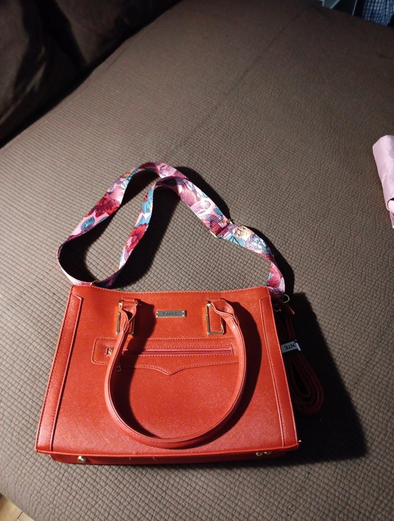 K&ouml;nnte beinhalten: Eine rote Ledertasche mit einem floralen Riemen. Die Tasche hat einen oberen Griff und einen Rei&szlig;verschluss.
