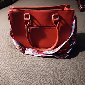 K&ouml;nnte beinhalten: Eine rote Ledertasche mit einem floralen Riemen. Die Tasche hat zwei obere Henkel und einen Rei&szlig;verschluss.