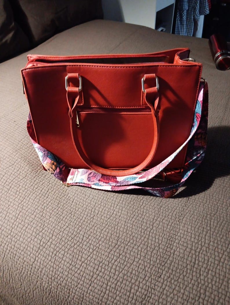 K&ouml;nnte beinhalten: Eine rote Ledertasche mit einem floralen Riemen. Die Tasche hat zwei obere Henkel und einen Rei&szlig;verschluss.