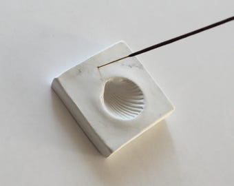 May include: A square, white incense holder with a seashell-shaped indentation. A thin, dark incense stick is placed in the holder. The holder is made of a smooth, matte material.