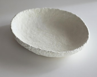 May include: A handmade, off-white oval bowl with a textured surface. The bowl has a rough, uneven edge and a shallow interior. The bowl is made of a paper-like material and is suitable for decorative purposes.