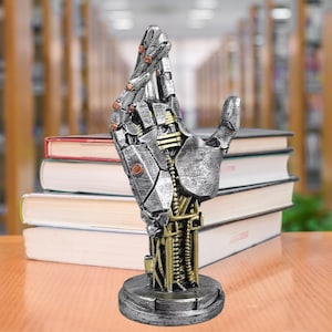 May include: A silver and gold robotic hand sculpture with a detailed design. The hand is raised with fingers extended and the palm facing forward. The sculpture is on a round base.