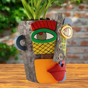 Pode incluir: Um vaso decorativo com um rosto abstrato. O corpo é cinza metálico texturizado. O rosto apresenta um olho vermelho e verde, um padrão de losangos amarelos, uma bochecha laranja e lábios vermelhos. Caules verdes de plantas emergem do topo.