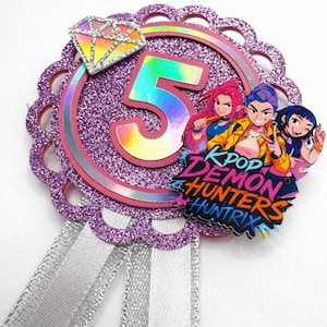 May include: A decorative badge with a glittery purple background, a large number 5 in rainbow colours, and a diamond-shaped accent. The badge features the text "KPOP DEMON HUNTRIX" and a silver ribbon.