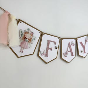 May include: A decorative banner spelling "FAY" with pink glitter letters on white pennant flags. The flags are edged in brown and strung on twine. One flag features a fairy illustration, others have butterflies and bees.