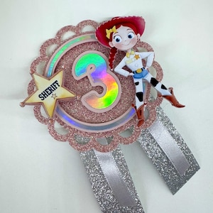 May include: A decorative badge featuring a rose gold glitter scalloped circle with a holographic number 3 in the centre. A yellow star with the word SHERIFF and a cutout of a cowgirl character are attached. Two silver glitter ribbons hang below.