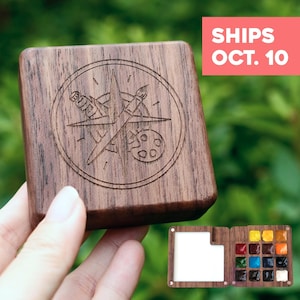 Pode incluir: Uma caixa de madeira quadrada de cor castanho-escuro para aguarelas com um design de bússola gravado a laser, incluindo um pincel e uma paleta. A caixa abre-se para revelar uma área de mistura branca e doze pequenas godés de aguarelas.
