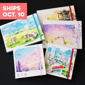 May include: Five rectangular sketchbooks featuring watercolor illustrations of a cow in a field, a traditional building, a starry path, a sunset landscape, and a city train scene. Each sketchbook is secured with a colorful elastic band closure.