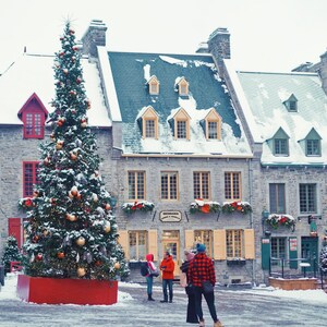 以下が含まれることがあります： 雪景色の中、石造りの建物の前に大きなクリスマスツリーが飾られています。建物には雪が積もった屋根と赤いアクセントがあります。雪が積もった広場を歩く人々。