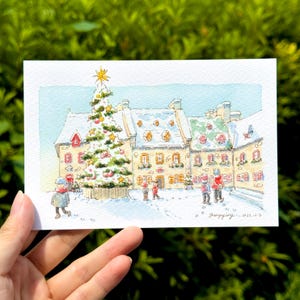 以下が含まれることがあります： 雪の降るクリスマスの風景を描いた水彩画。雪に覆われた屋根の建物前に、装飾されたクリスマスツリーが立っています。雪の中を歩く人々の姿。白いカードに描かれたアートワークです。