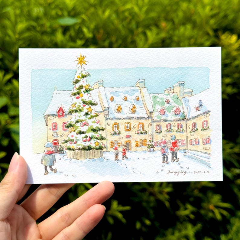 以下が含まれることがあります： 雪の降るクリスマスの風景を描いた水彩画。雪に覆われた屋根の建物前に、装飾されたクリスマスツリーが立っています。雪の中を歩く人々の姿。白いカードに描かれたアートワークです。