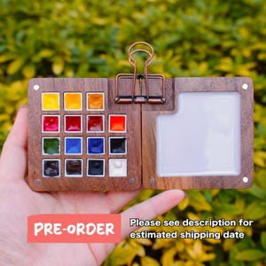 May include: A wooden watercolor paint palette with 16 colorful paint wells and a mixing area. The palette is held open with a copper-colored clip. The text "PRE-ORDER" is visible on the bottom left. The text "Please see description for estimated shipping date" is on the bottom right.
