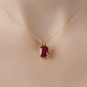 Puede incluir: Un collar de cadena de oro con un colgante de piedra roja. El colgante tiene forma cuadrada con esquinas redondeadas.