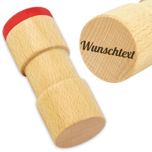 Könnte beinhalten: Ein Holzstempel mit roter Oberseite und dem Text "Wunschtext" auf der Seite. Der Stempel besteht aus hellem Holz und hat eine zylindrische Form. Der Stempel ist zum Aufdrucken von Text vorgesehen.