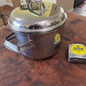 May include: A stainless steel cooking pot with a lid and handle, next to a 16-foot Stanley Powerlock II measuring tape. The pot has a shiny, reflective surface. The measuring tape is yellow and gray.