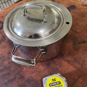 May include: A stainless steel pot with a lid and handle, and a measuring tape. The pot is round with a handle on the side and a handle on the lid. The measuring tape is yellow and has black text that reads "16' STANLEY POWERLOCK II".
