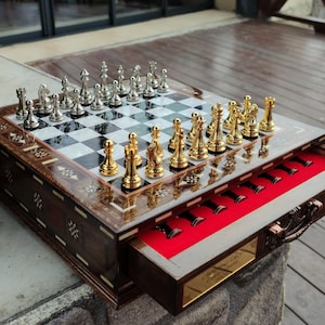 K&ouml;nnte beinhalten: Ein Schachbrett aus Holz mit einer Schublade zum Verstauen der Figuren. Das Brett hat ein schwarz-wei&szlig;es Schachbrettmuster und ist mit aufwendigen Designs verziert. Die Schachfiguren sind aus Metall und in Gold und Silber gehalten.