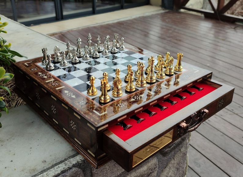 K&ouml;nnte beinhalten: Ein Schachbrett aus Holz mit einer Schublade zum Verstauen der Figuren. Das Brett hat ein schwarz-wei&szlig;es Schachbrettmuster und ist mit aufwendigen Designs verziert. Die Schachfiguren sind aus Metall und in Gold und Silber gehalten.