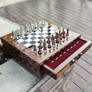K&ouml;nnte beinhalten: Ein Schachbrett aus Holz mit einer Schublade zum Verstauen der Figuren. Das Brett hat ein schwarz-wei&szlig;es Schachbrettmuster und ist mit aufwendigen Blumendesigns verziert. Die Schachfiguren sind aus Metall und in der Startposition auf dem Brett angeordnet.