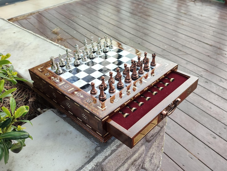 K&ouml;nnte beinhalten: Ein Schachbrett aus Holz mit einer Schublade zum Verstauen der Figuren. Das Brett hat ein schwarz-wei&szlig;es Schachbrettmuster und ist mit aufwendigen Blumendesigns verziert. Die Schachfiguren sind aus Metall und in der Startposition auf dem Brett angeordnet.