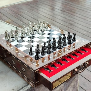 K&ouml;nnte beinhalten: Ein Schachspiel aus Holz mit einer Klappbox. Das Schachbrett ist mit Perlmutt eingelegt und die Figuren sind silber und schwarz. Die Box hat ein rotes Filz-Innenfutter und einen Messinggriff.